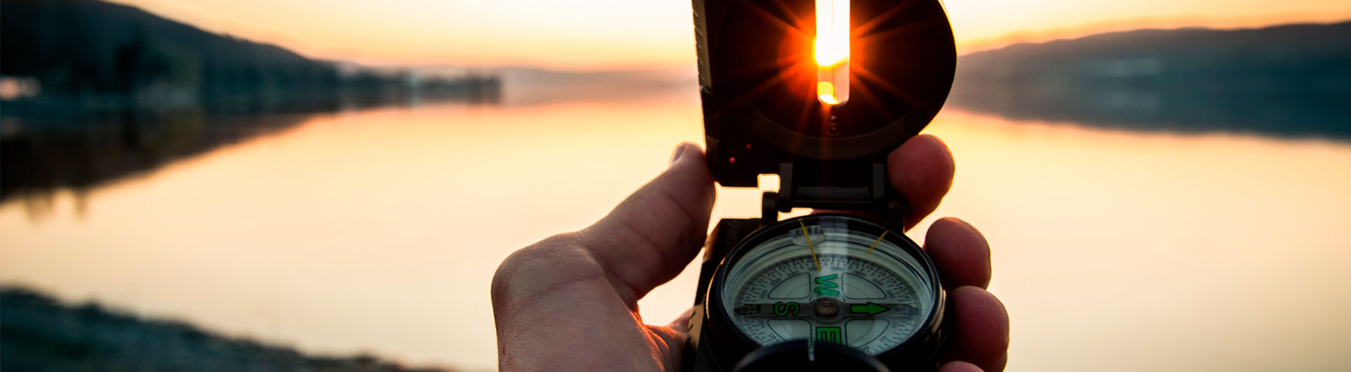Käsi pitelee kompassia joka osoittaa nousevan auringon valaisemaa järvimaisemaa kohti.