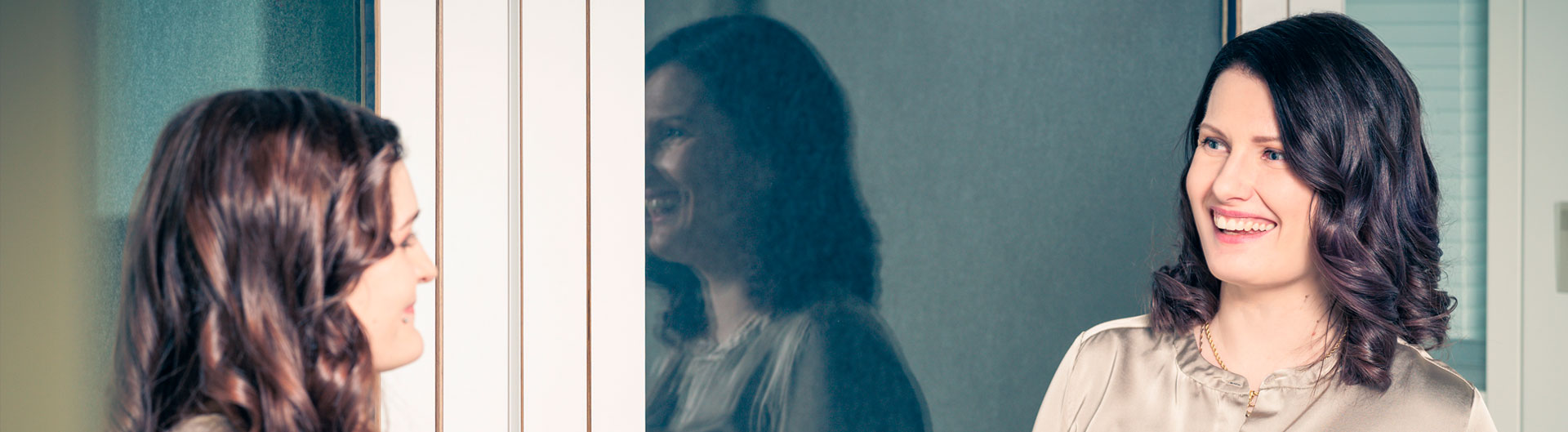 Kaksi hymyilevää pitkähiuksista naista keskustelee, taustalla lasiovet joista toisen naisen peilikuva heijastuu.
